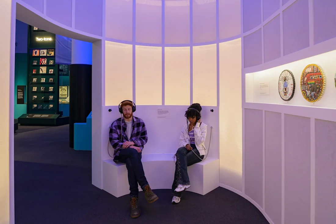 Two people sat in the Beyond the Bassline exhibition with headphones on.