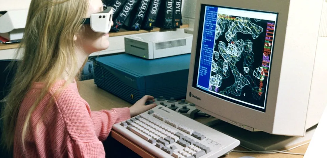 A girl with a VR headset on sat in front of a computer which shows 3D proteins