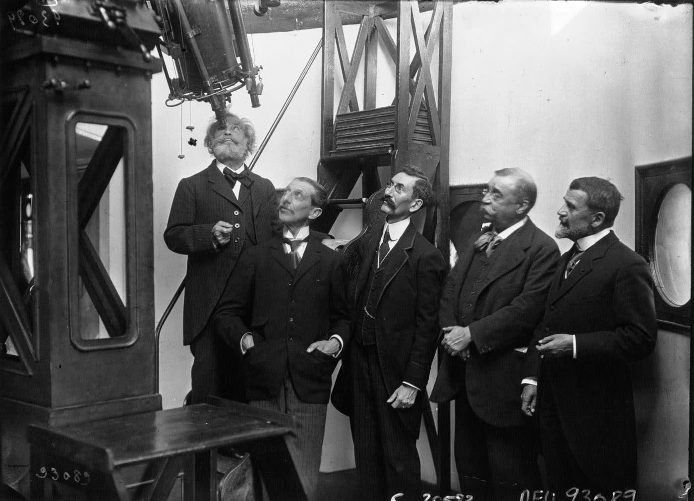 black and white photograph, a man looks through a large telescope while other men stand beside him.