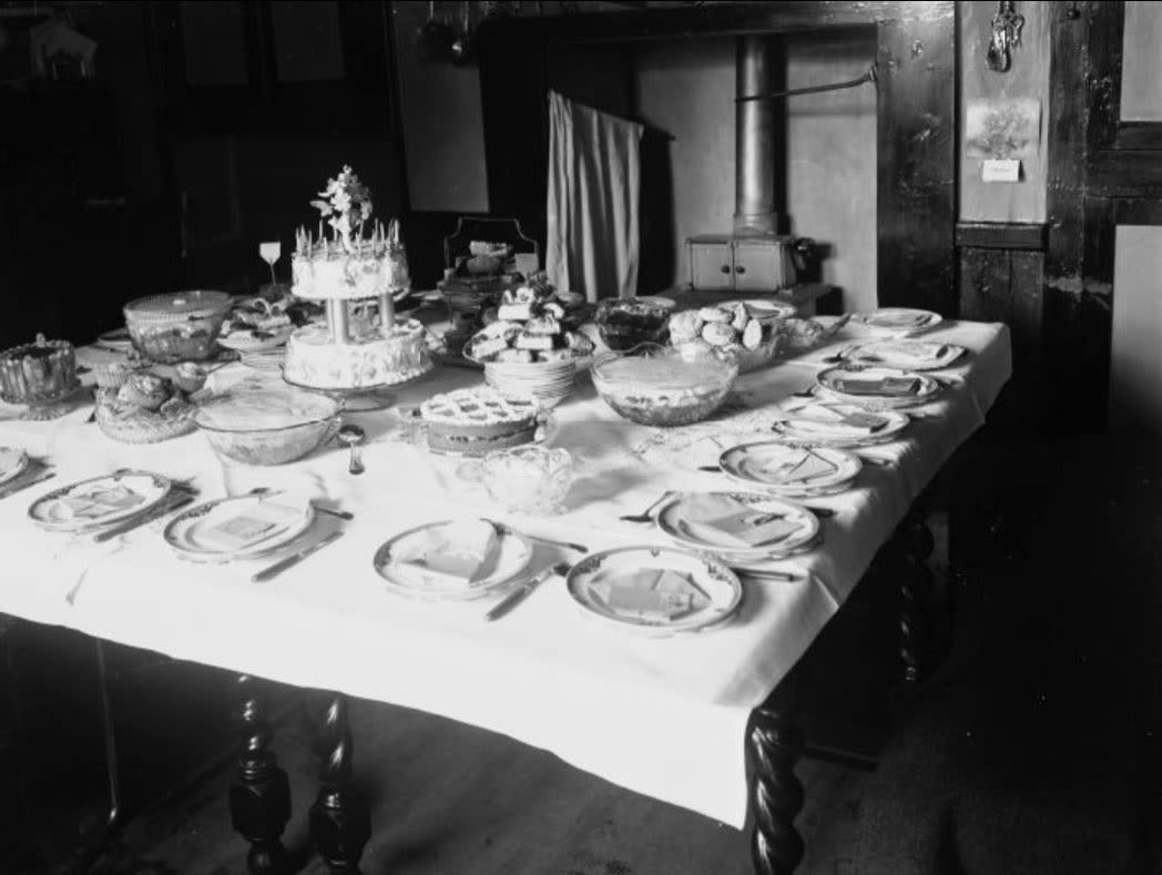 black and white photograph, a table covered in different food as well as plates and cutlery.