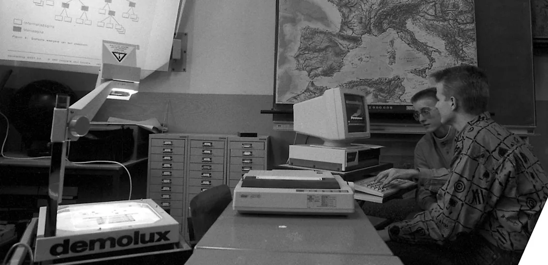 A photograph of two young people looking at a computer, in a room with maps on the wall and a projector. 
