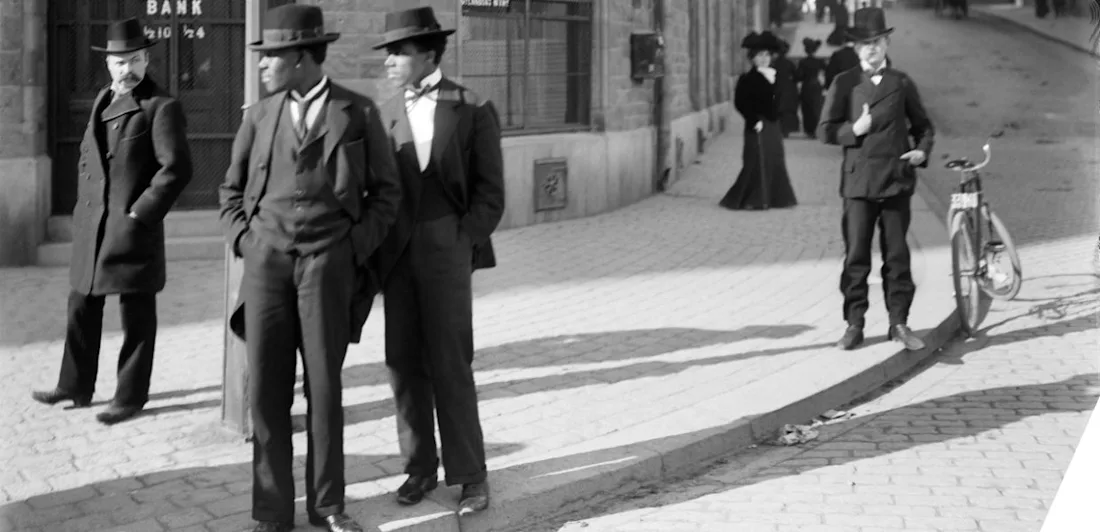 A black and white photo from 1901 depicting a street scene at Nyborplan in Stockholm. Two Black men are seen from the profile standing on the street, a passerby looking at them. A bank building, a bicycle and more people are visible in the background.