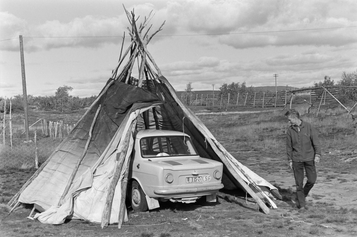 black and white photograph, a man standing by a car which is parked underneath a tent.