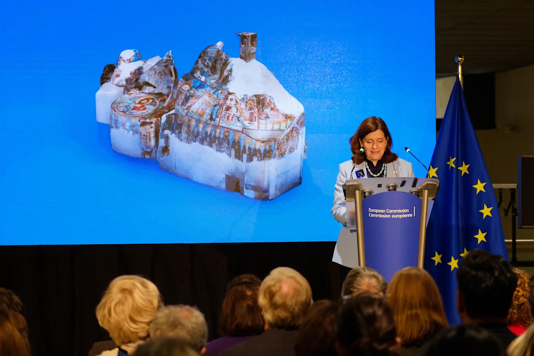 A woman speaks at a podium near an EU flag; a crowd listens and a model building is displayed on a screen behind her.