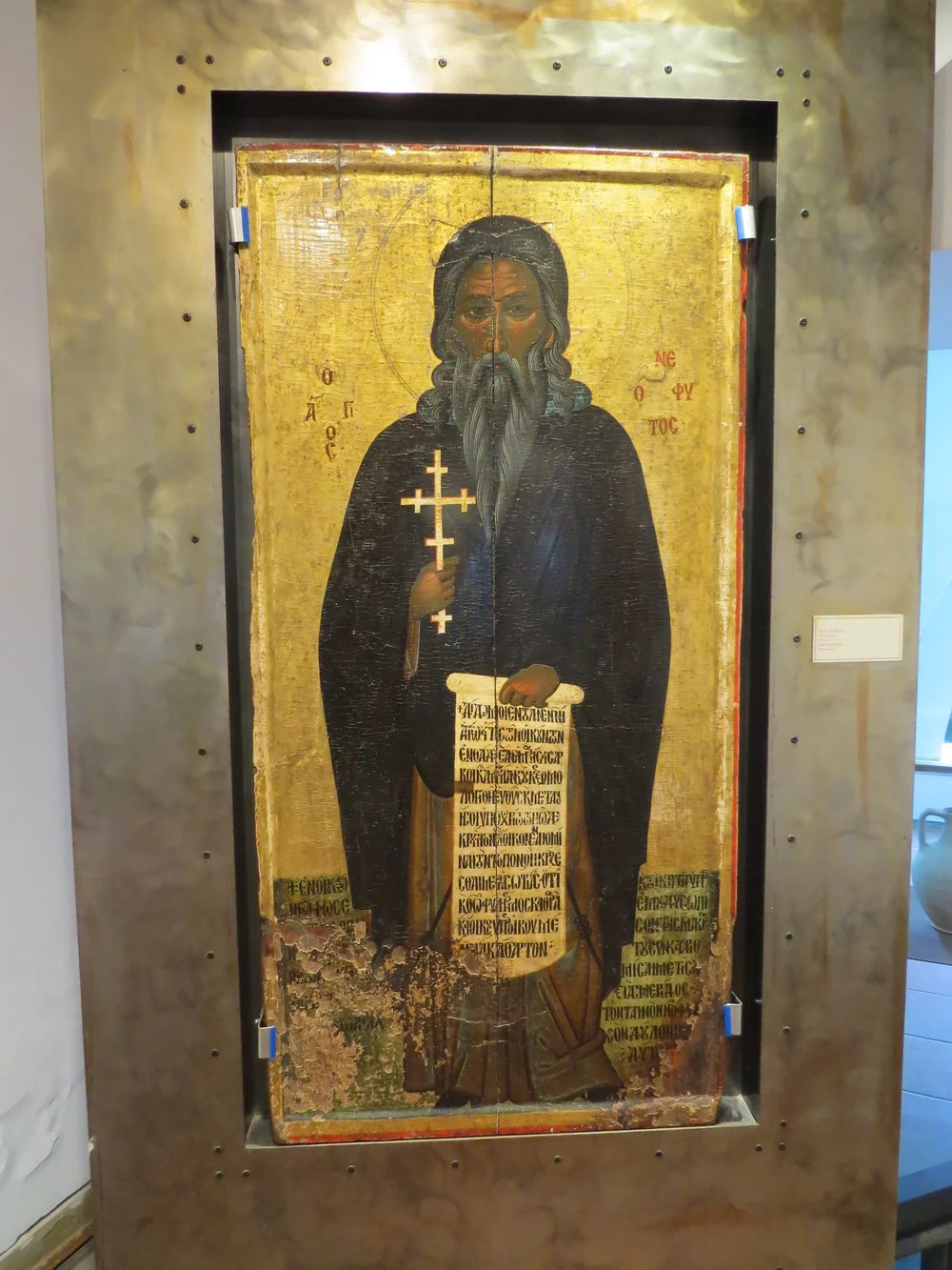 A large, 15th century icon, made of egg tempera and gold on wood, showing St. Neophytos with grey hair and a beard, dressed in monastic garments, holding a cross and displaying an inscribed scroll. He is standing on a green ground inscribed with a dense text in calligraphic Greek majuscule, pertaining to the dedication of the icon. The name of the saint is written with red letters in Greek majuscule on the gold background, the latter extending on the raised border with a red band running along its edge.
The icon is exhibited in the museum of the Monastery of St. Neophytos, which is opposite the Enkleistra.