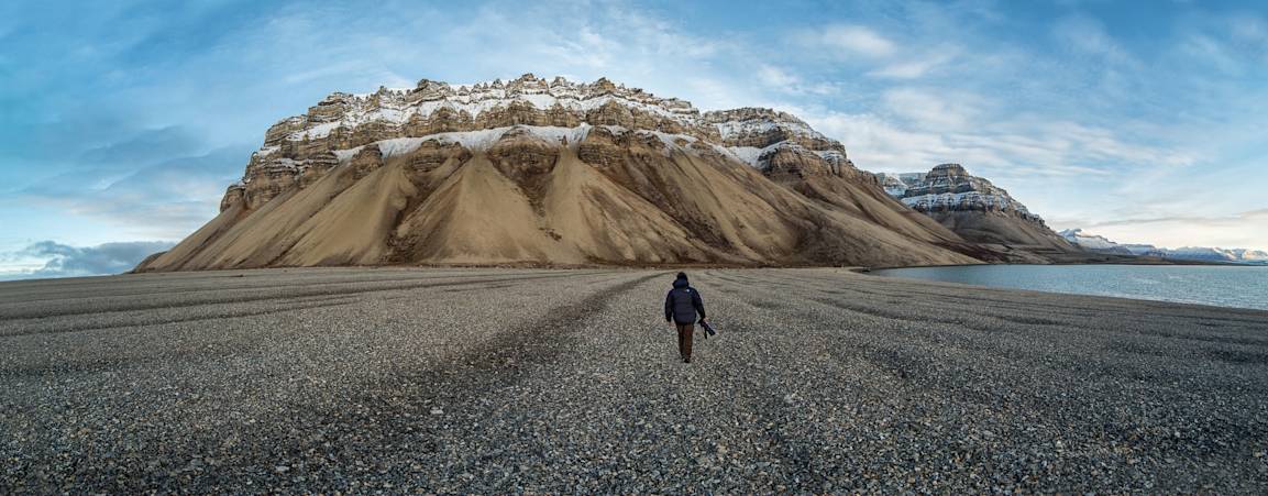 A photographer walks along a rocky beach with mountains in the background in Svalbard during summer