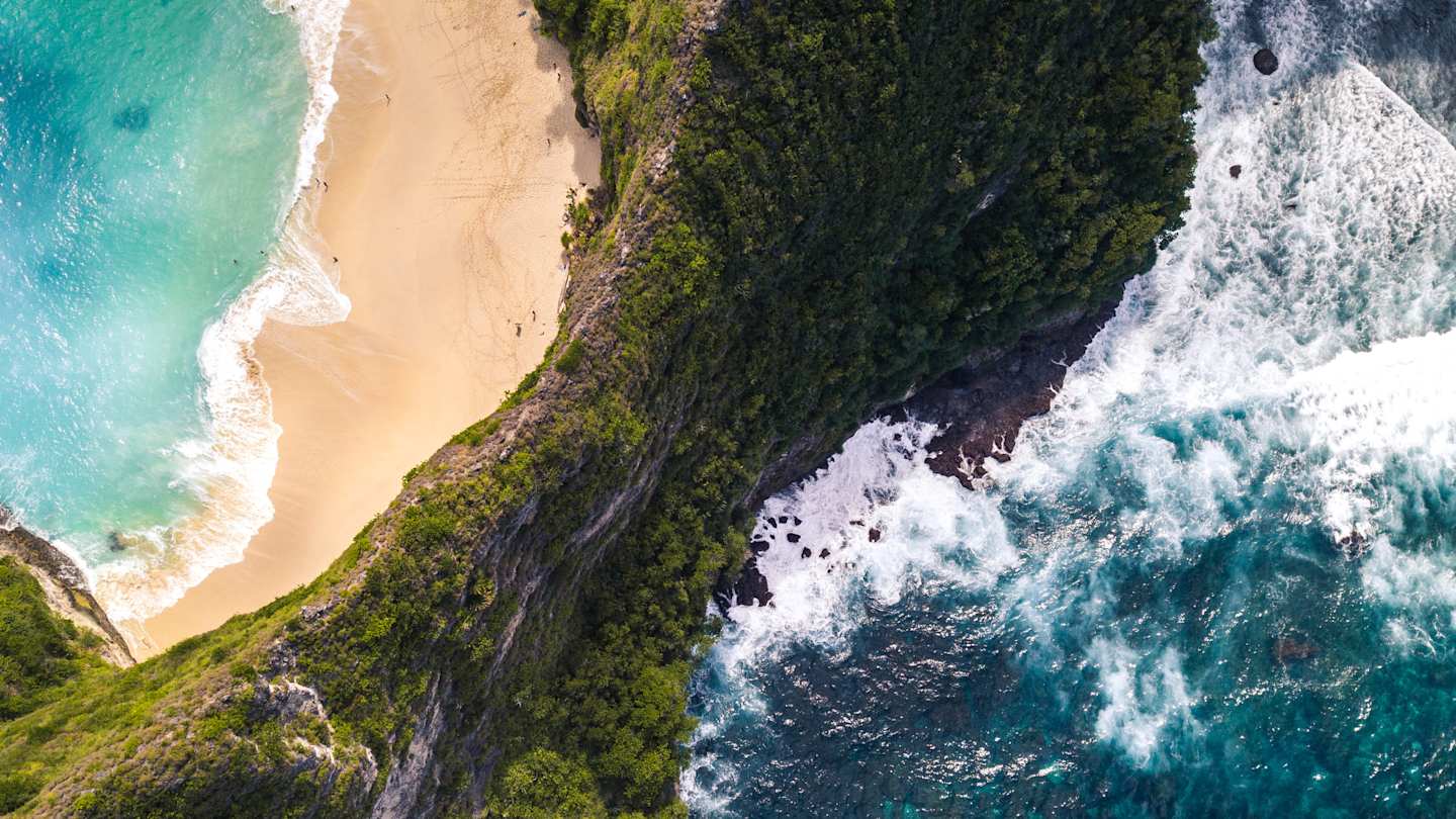 A bird's eye view of the green cliffs, golden sand, and crystalline blue water at Kelingking Beach, Indonesia