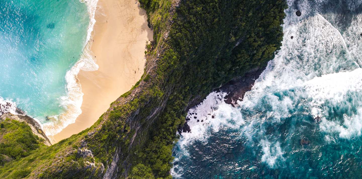 A bird's eye view of the green cliffs, golden sand, and crystalline blue water at Kelingking Beach, Indonesia