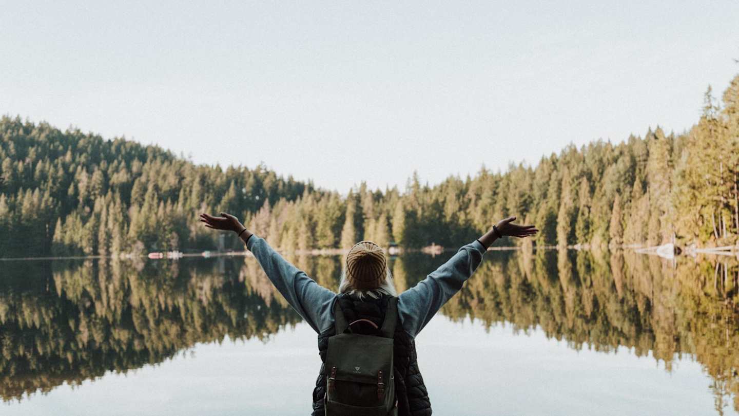 A woman standing by a large lake a green trees in a happy pose