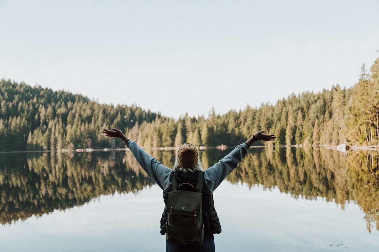A woman standing by a large lake a green trees in a happy pose