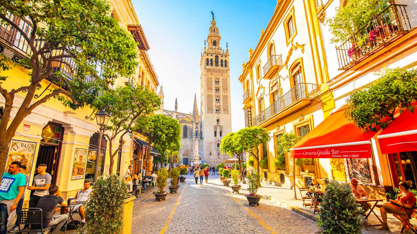 Sunny, cobbled street lined with trees and eateries, with views of Giralda Tower, Seville