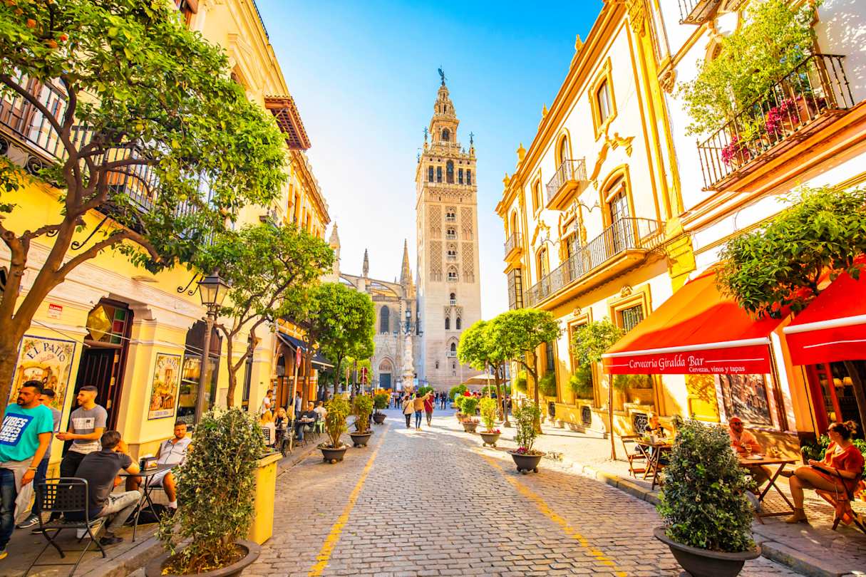 Sunny, cobbled street lined with trees and eateries, with views of Giralda Tower, Seville