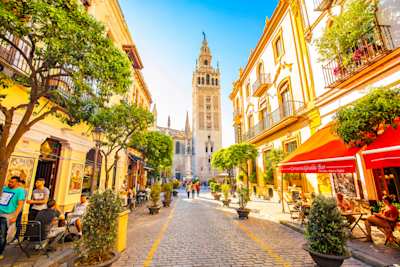 Sunny, cobbled street lined with trees and eateries, with views of Giralda Tower, Seville