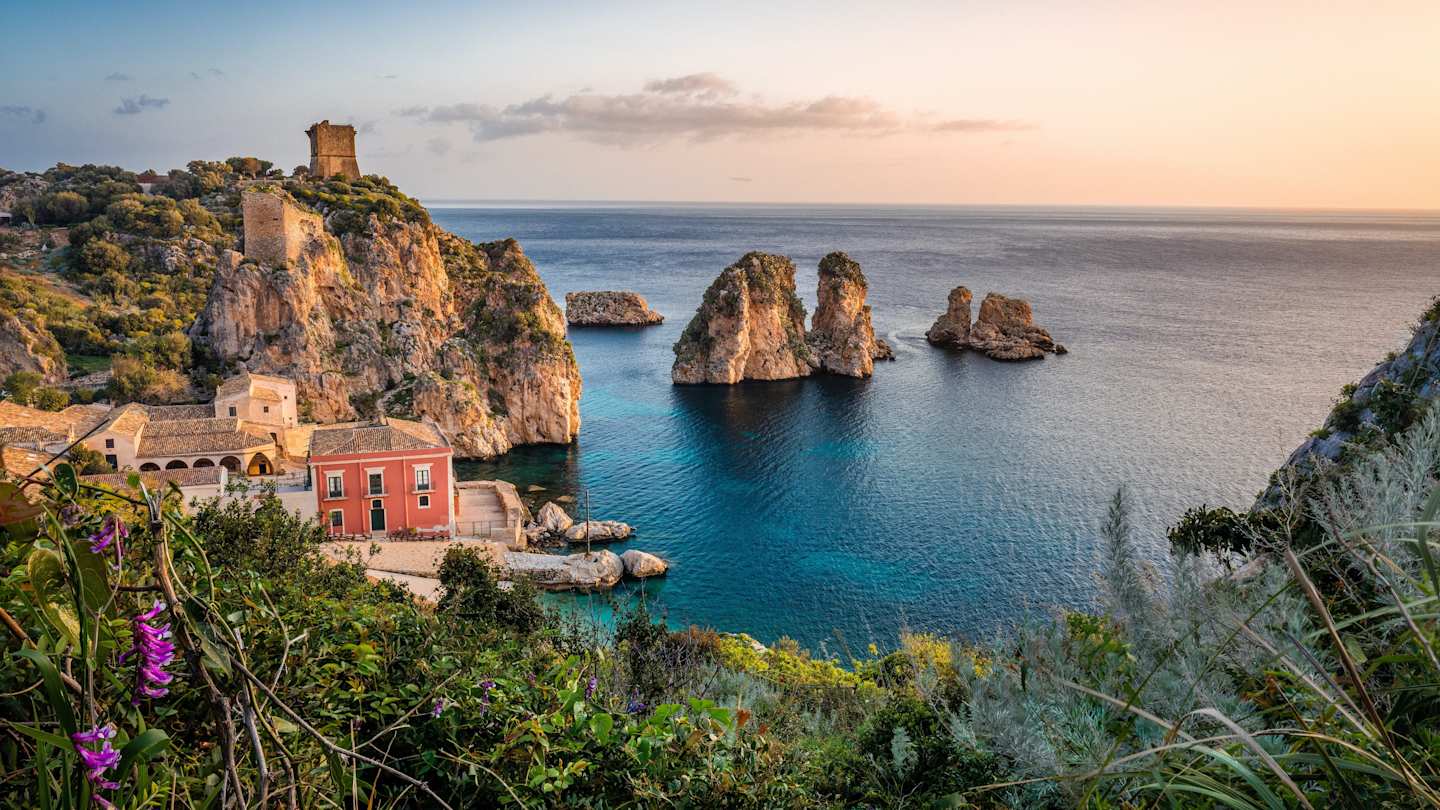 A small red house next to the blue sea and large rocks in Sicily, Italy