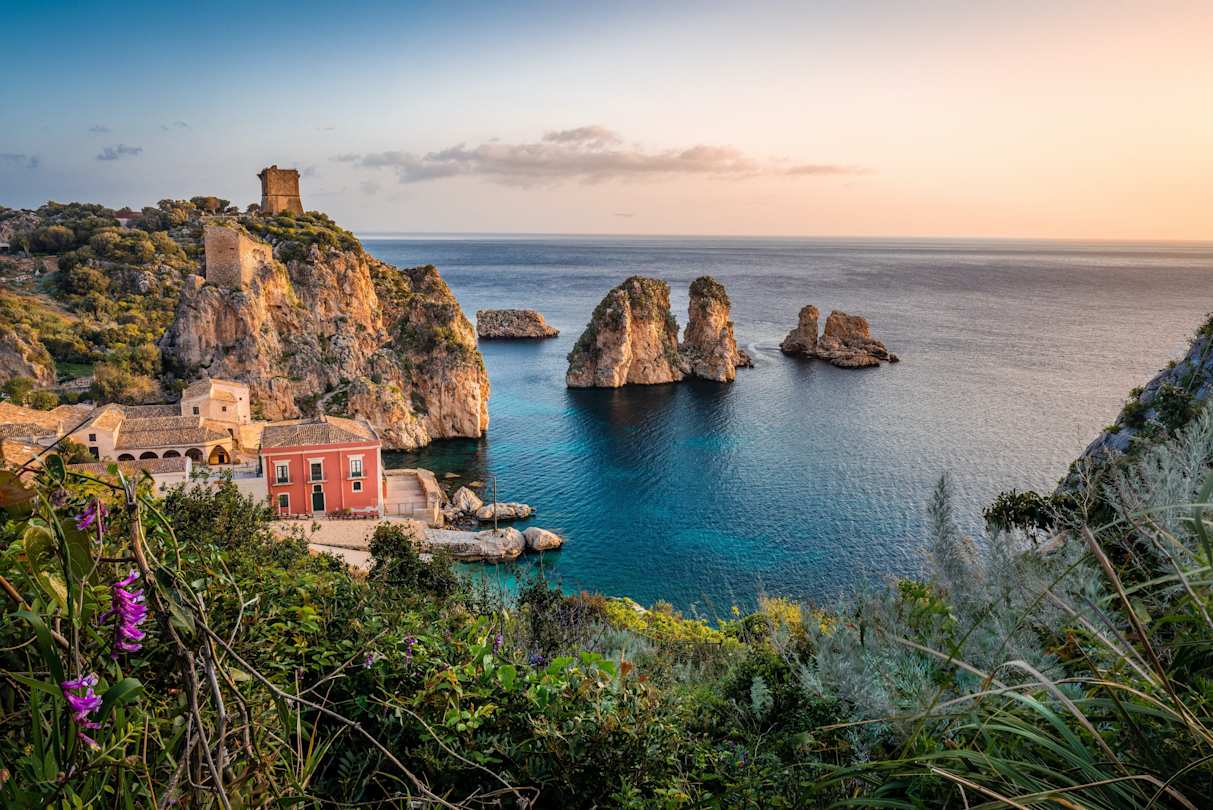 A small red house next to the blue sea and large rocks in Sicily, Italy