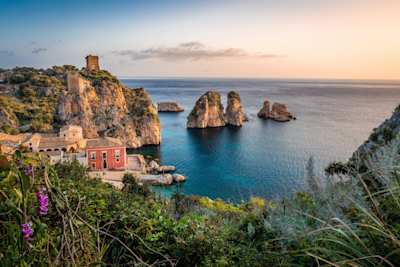 A small red house next to the blue sea and large rocks in Sicily, Italy