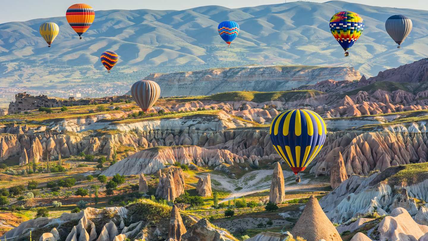 Multiple colourful hot air balloons over green fields and jagged rocks in a fairytale-like setting, Cappadocia, Turkey