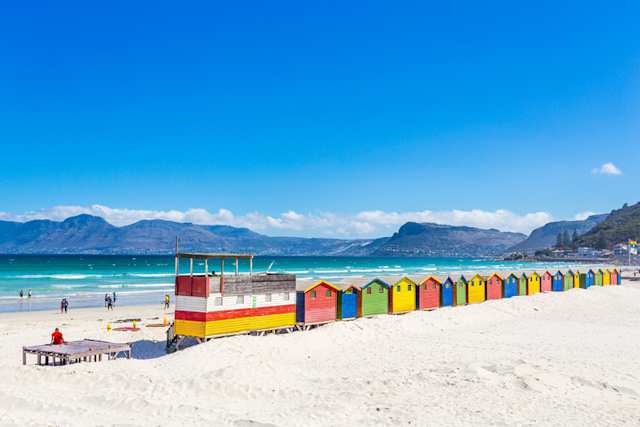 Colourful beach shacks on Muizenberg Beach contrasted with white sand and blue ocean, mountains in the distance, Cape Town