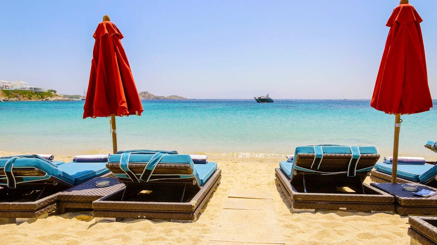 Sun loungers and red parasols looking out to sea from the soft sand on a beach in Mykonos, Greece