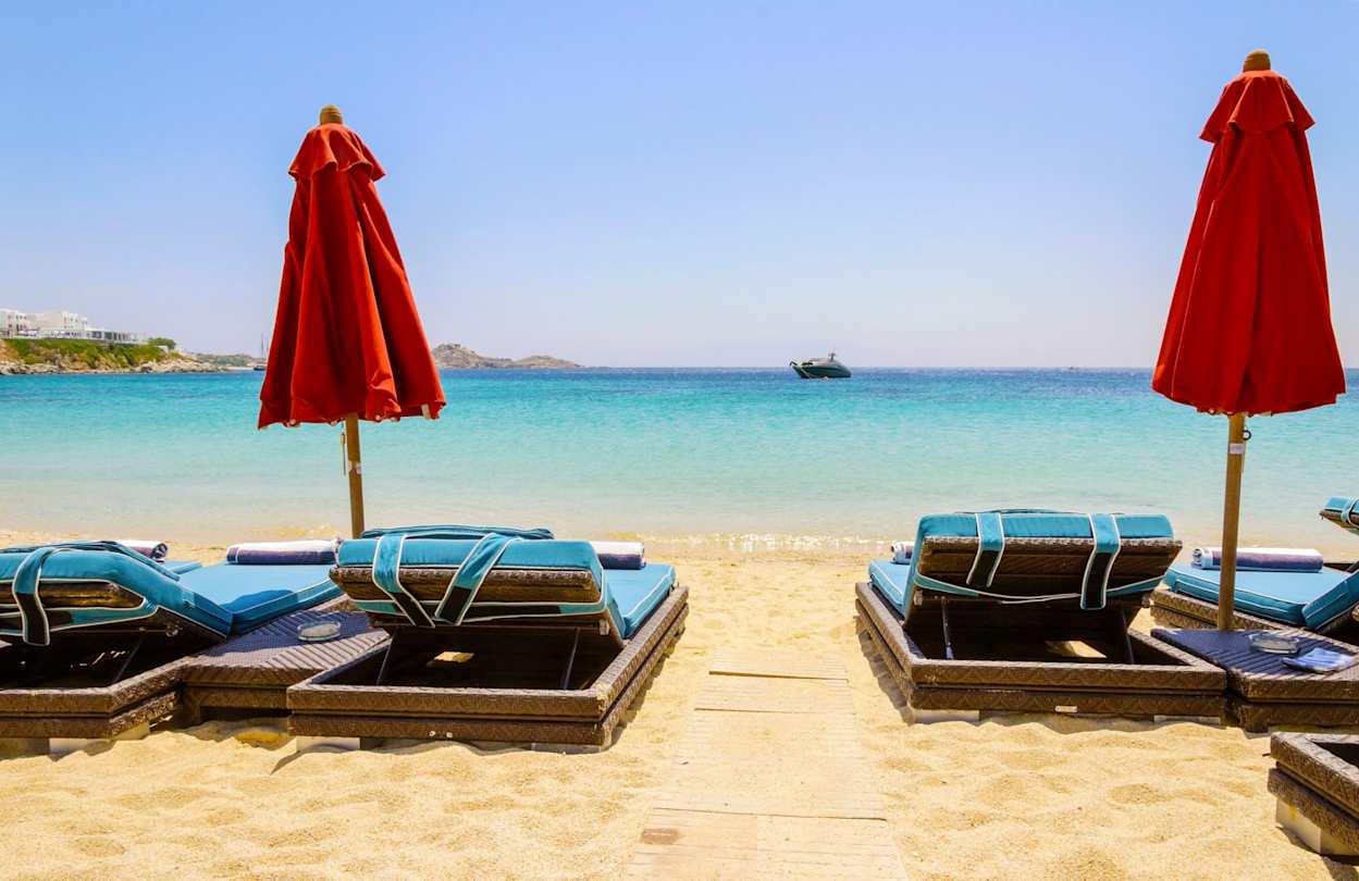 Sun loungers and red parasols looking out to sea from the soft sand on a beach in Mykonos, Greece