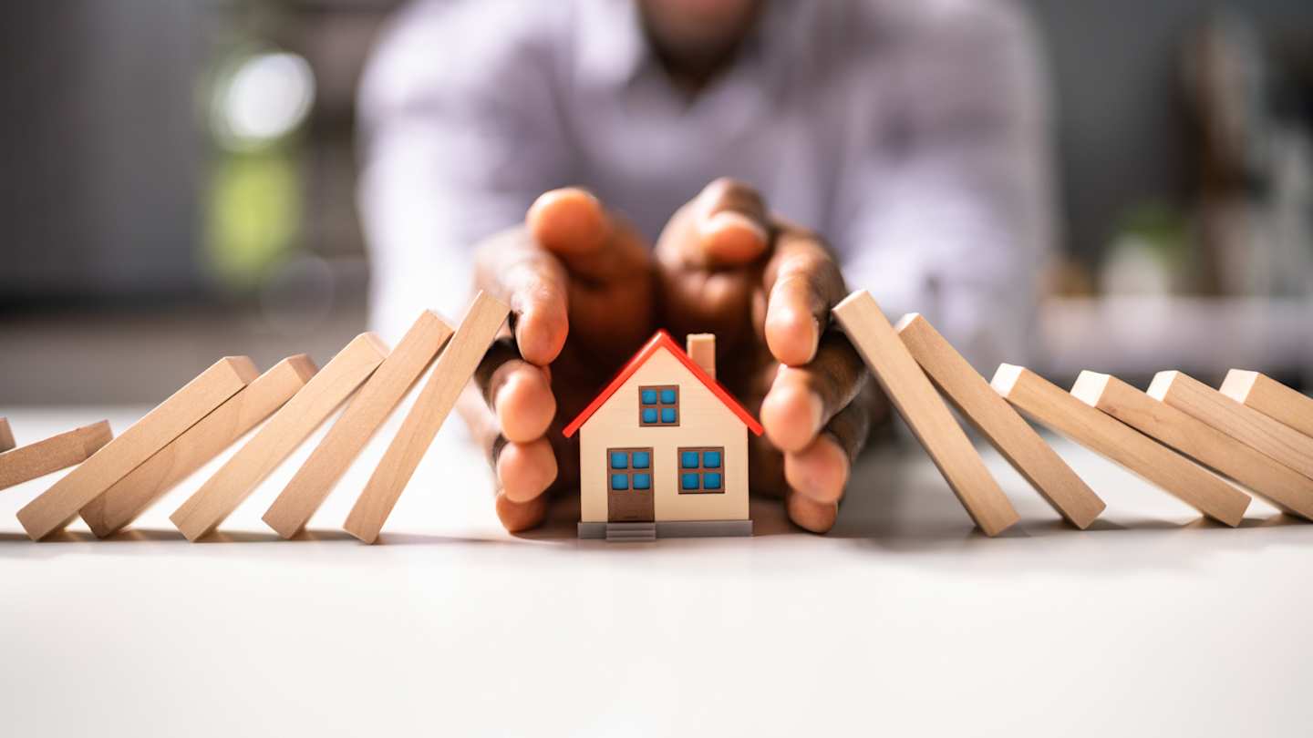 A man protecting a small model house from dominoes to represent holiday home insurance