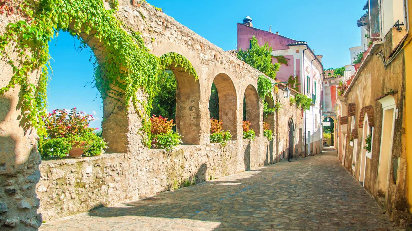 Old stone wall with arches, vines and flowers on ancient European street, Ravello, near Amalfi