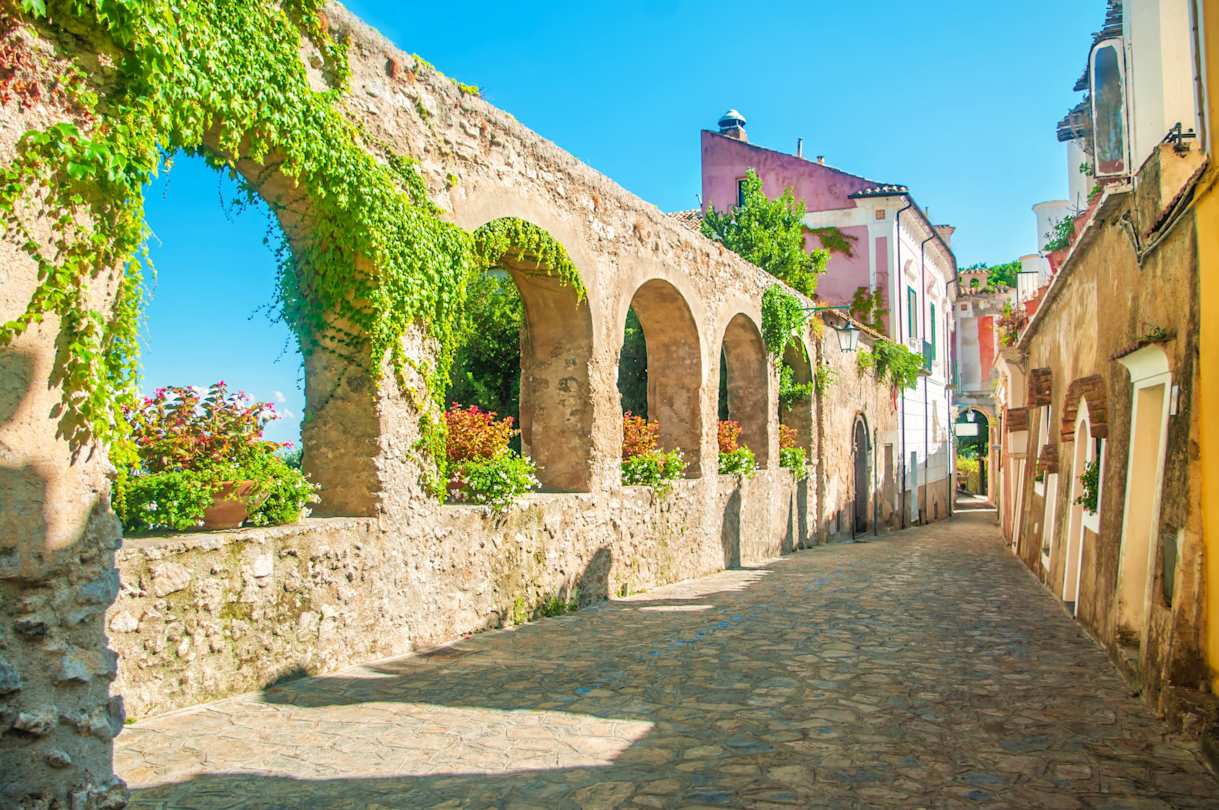 Old stone wall with arches, vines and flowers on ancient European street, Ravello, near Amalfi