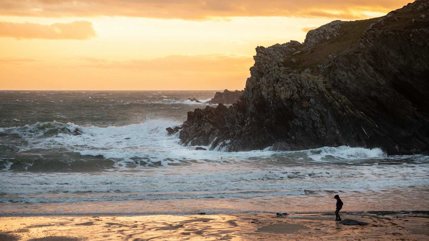 A person on the sand in front of a large rock in the sea on a beach at sunset, Anglesey, Wales, UK