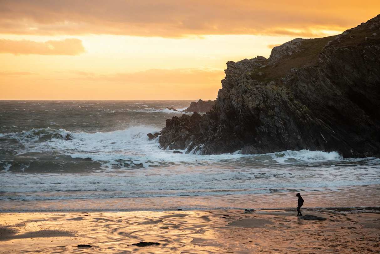 A person on the sand in front of a large rock in the sea on a beach at sunset, Anglesey, Wales, UK