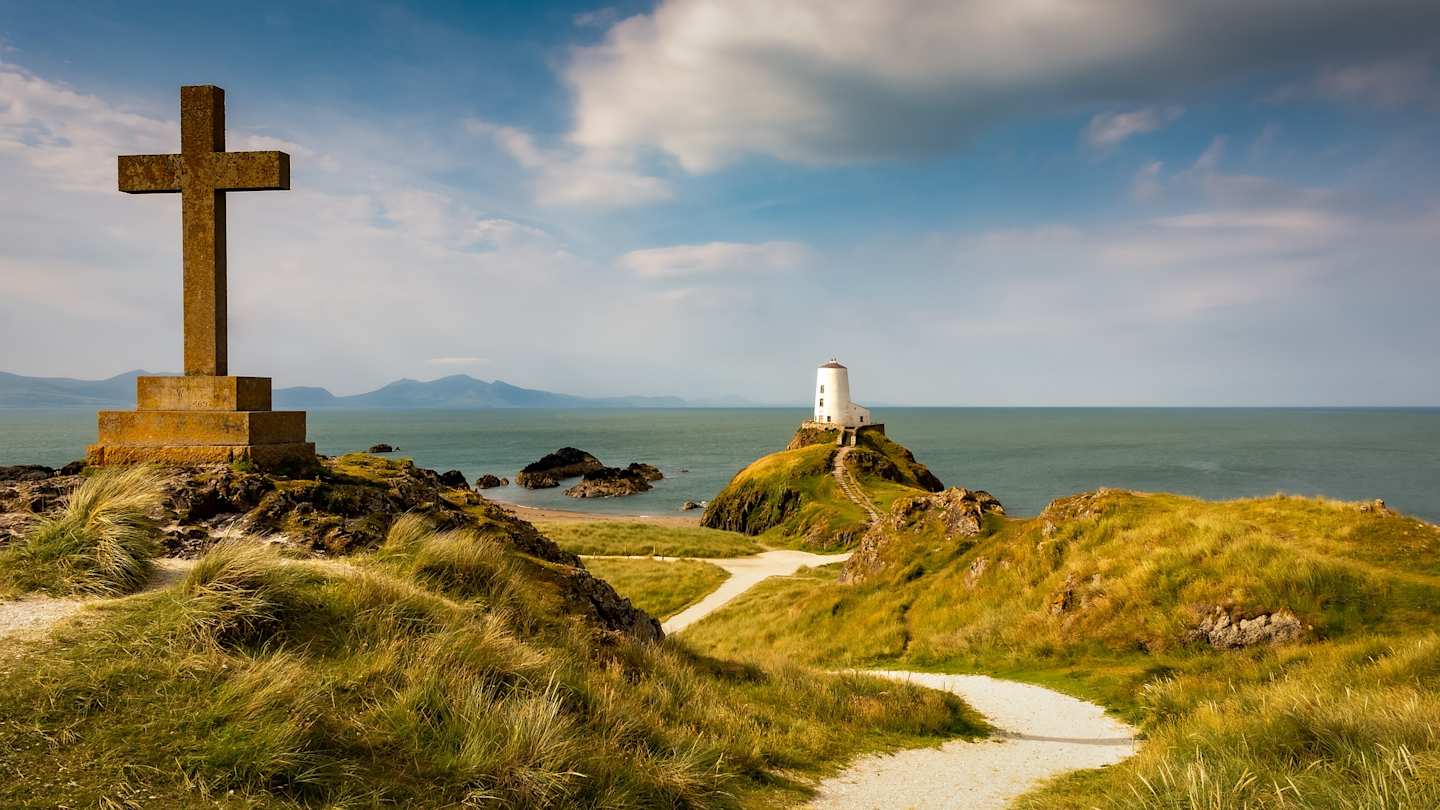 A brown tombstone and a white lighthouse along a coastal path in Llanddwyn Island overlooking the sea, Anglesey, Wales, UK