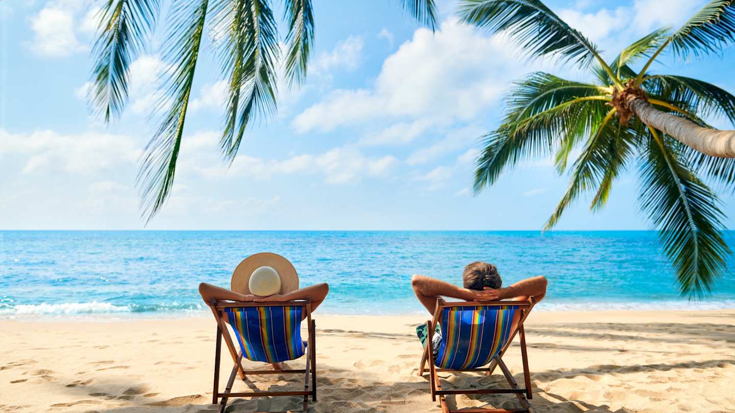 A couple relaxing on sun loungers on a beach by palm trees looking out to sea in a sunny location