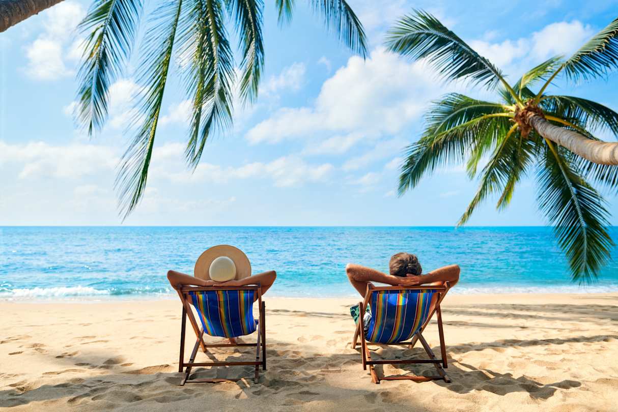 A couple relaxing on sun loungers on a beach by palm trees looking out to sea in a sunny location