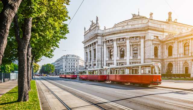 A red and white tram in front of a large theatre on a sunny day in Vienna, Austria