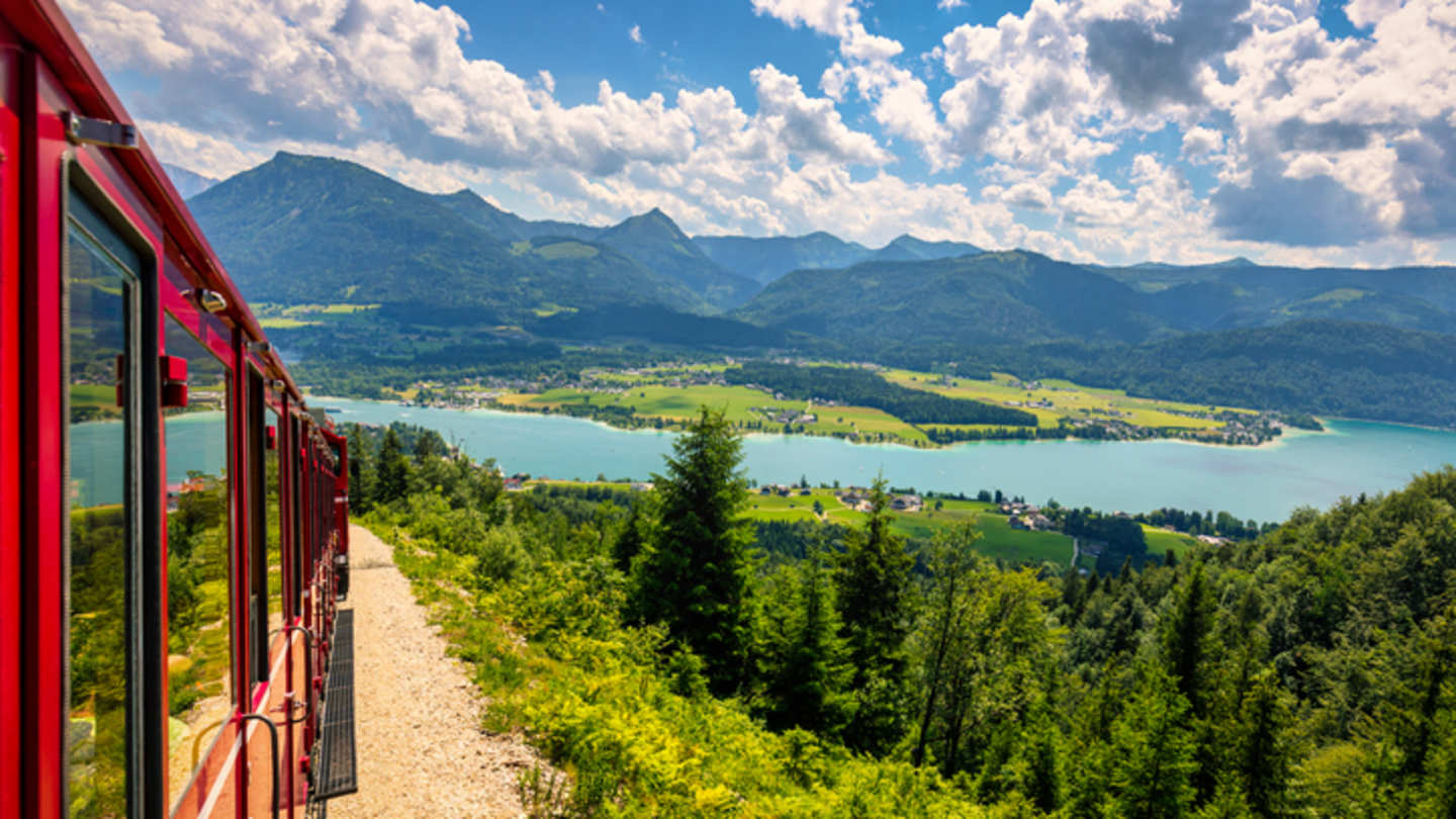 A red train driving through green hills by a lake in Salzburg, Austria