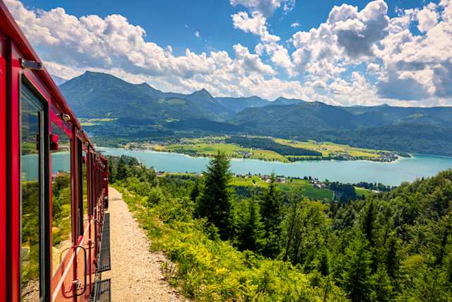 A red train driving through tall green hills by a large lake, Salzburg, Austria
