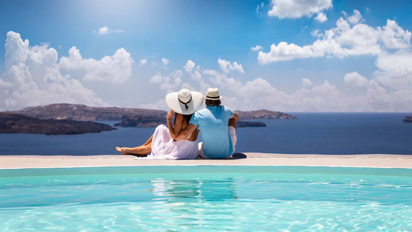 A young couple sitting by a swimming pool looking out into the sea, Santorini, Greece