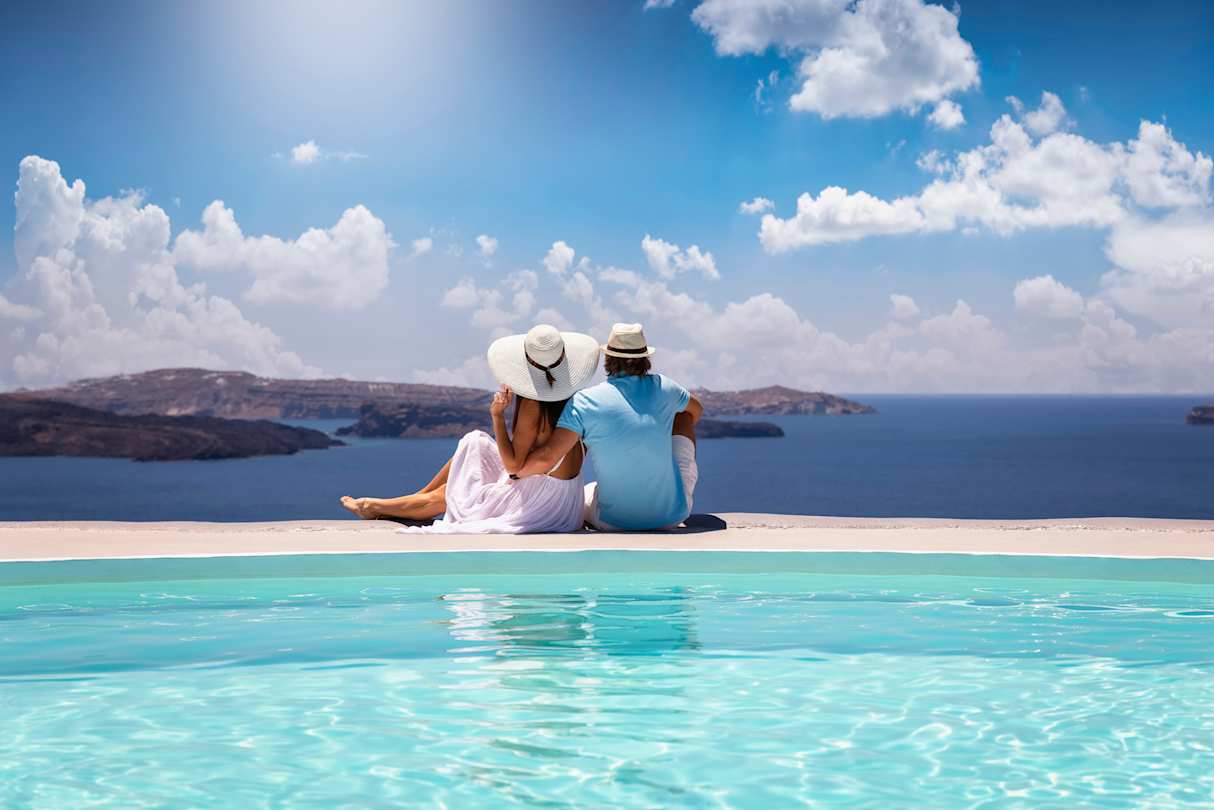 A young couple sitting by a swimming pool looking out into the sea, Santorini, Greece