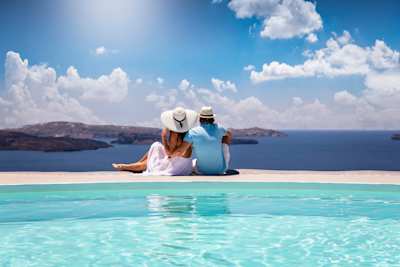 A young couple sitting by a swimming pool looking out into the sea, Santorini, Greece