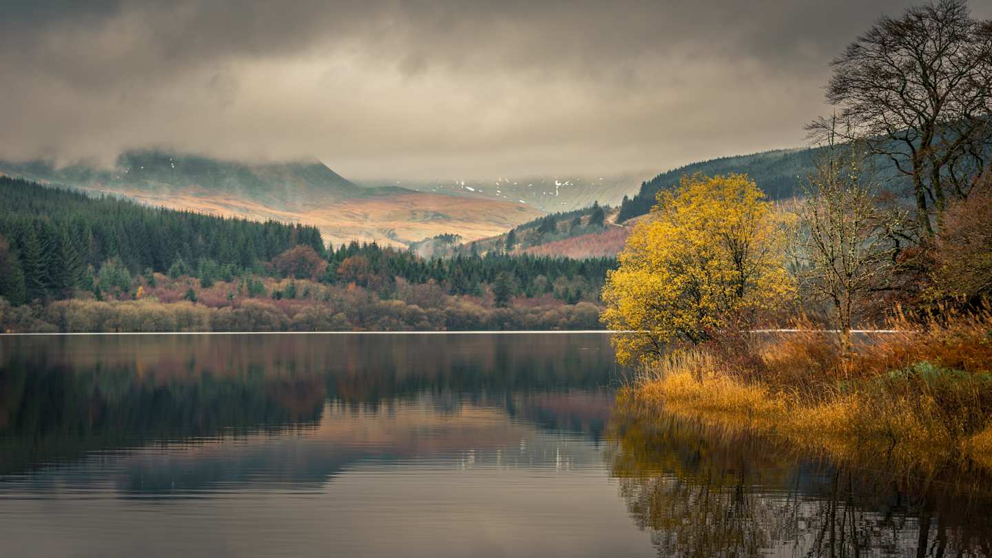A still lake in front of trees and mountains at Brecon Beacons, Wales, UK