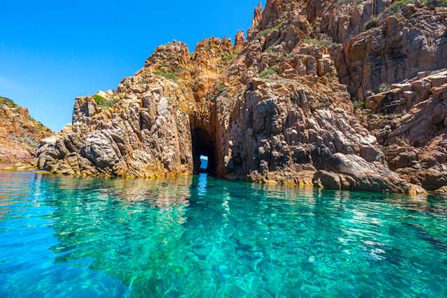 A view of clear blue water with large brown rocks and an arch, Calanches de Piana, Corsica, France