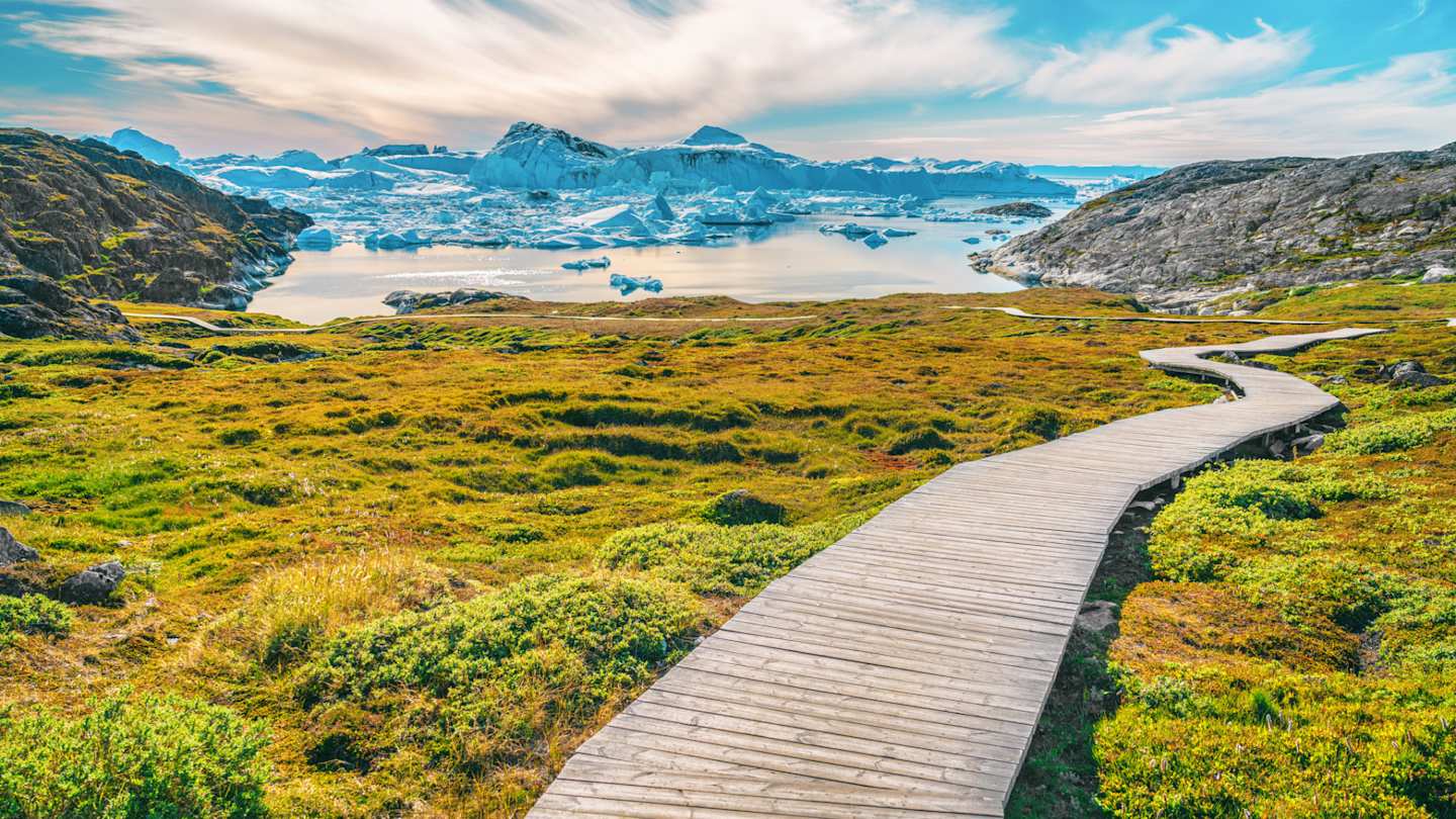 A long, empty boardwalk in a large green field in front of distant snow-capped mountains, Ilulissat Icefjord, Greenland