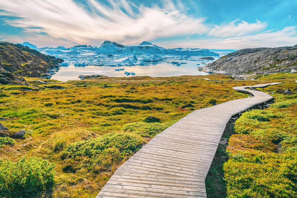 A long, empty boardwalk in a large green field in front of distant snow-capped mountains, Ilulissat Icefjord, Greenland