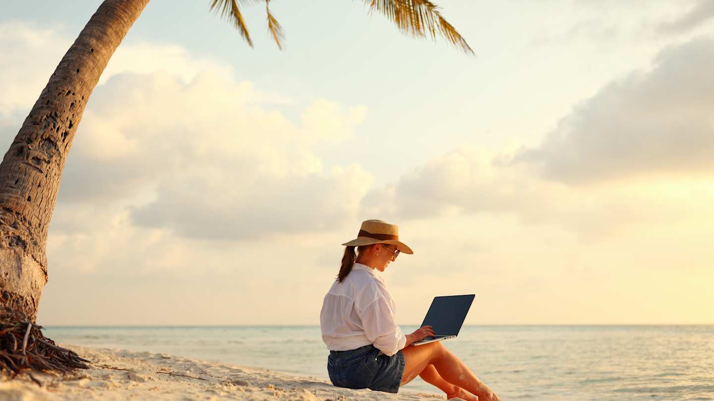 A digital nomad sitting on a sandy beach with her laptop under a palm tree