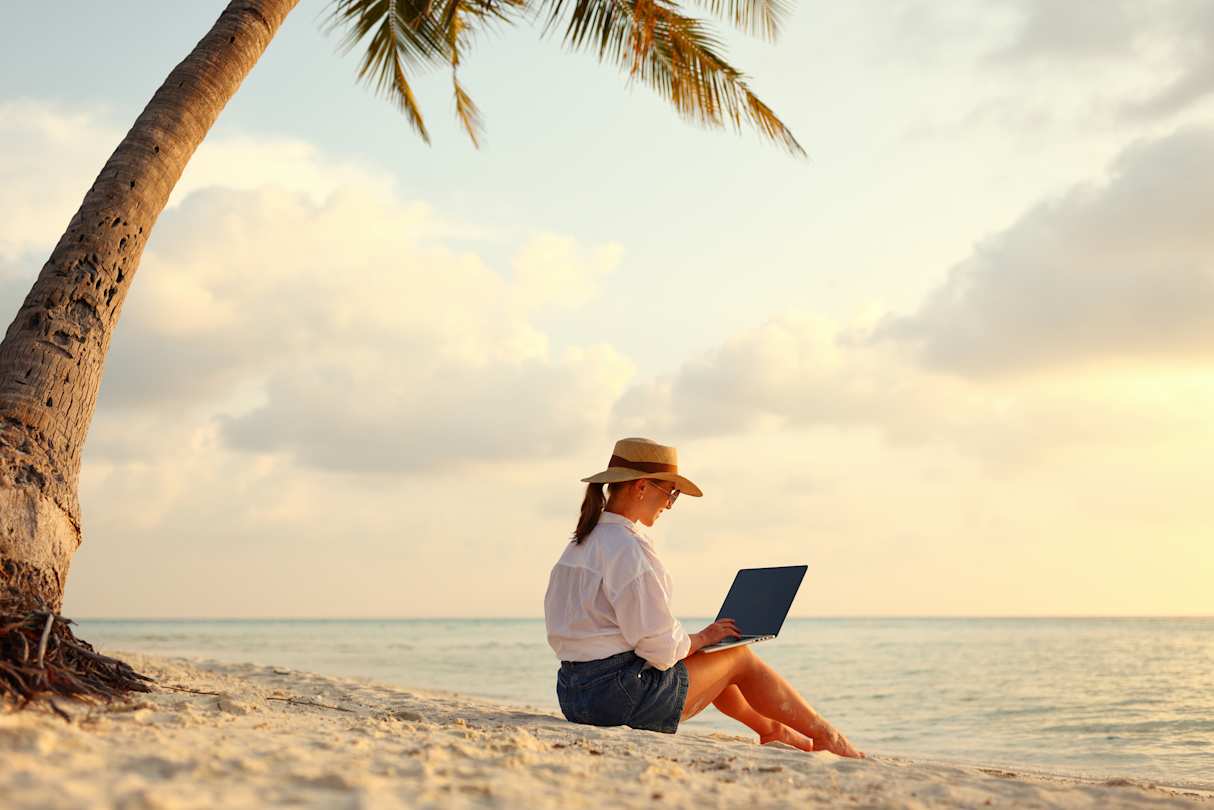 A digital nomad sitting on a sandy beach with her laptop under a palm tree