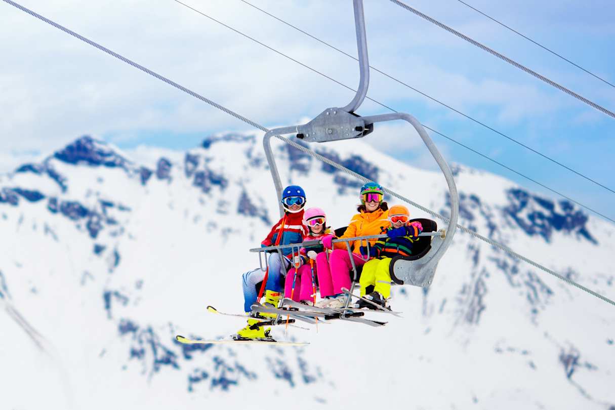 A family wearing colourful clothes on a ski lift at a ski resort in front of snowy mountains