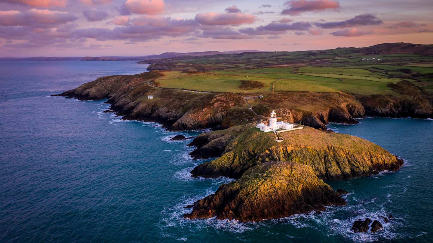 Aerial view of Strumble Head Lighthouse at dusk, with purple and pink sky and rocky coastline, Pembrokeshire