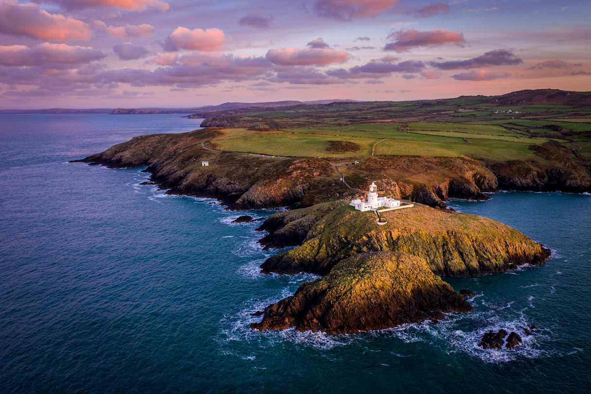 Aerial view of Strumble Head Lighthouse at dusk, with purple and pink sky and rocky coastline, Pembrokeshire