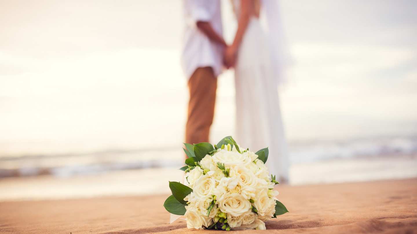 A close up view of a white bouquet of flowers in front of a newly-married couple at their beach wedding