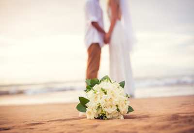 A close up view of a white bouquet of flowers in front of a newly-married couple at their beach wedding