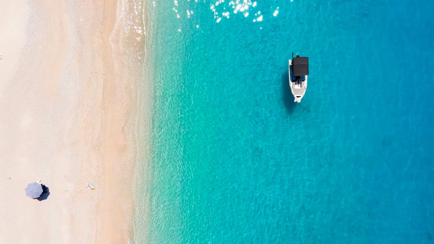 A bird's eye view of one parasol on the sand and a small boat in the clear blue sea at a beach in Kefalonia, Greece